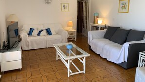 Sala de estar com dois sofás brancos, uma mesa de centro de vidro, uma televisão e um armário branco.