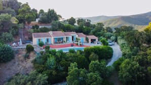 Uma casa grande com piscina, rodeada por vegetação exuberante e colinas.
