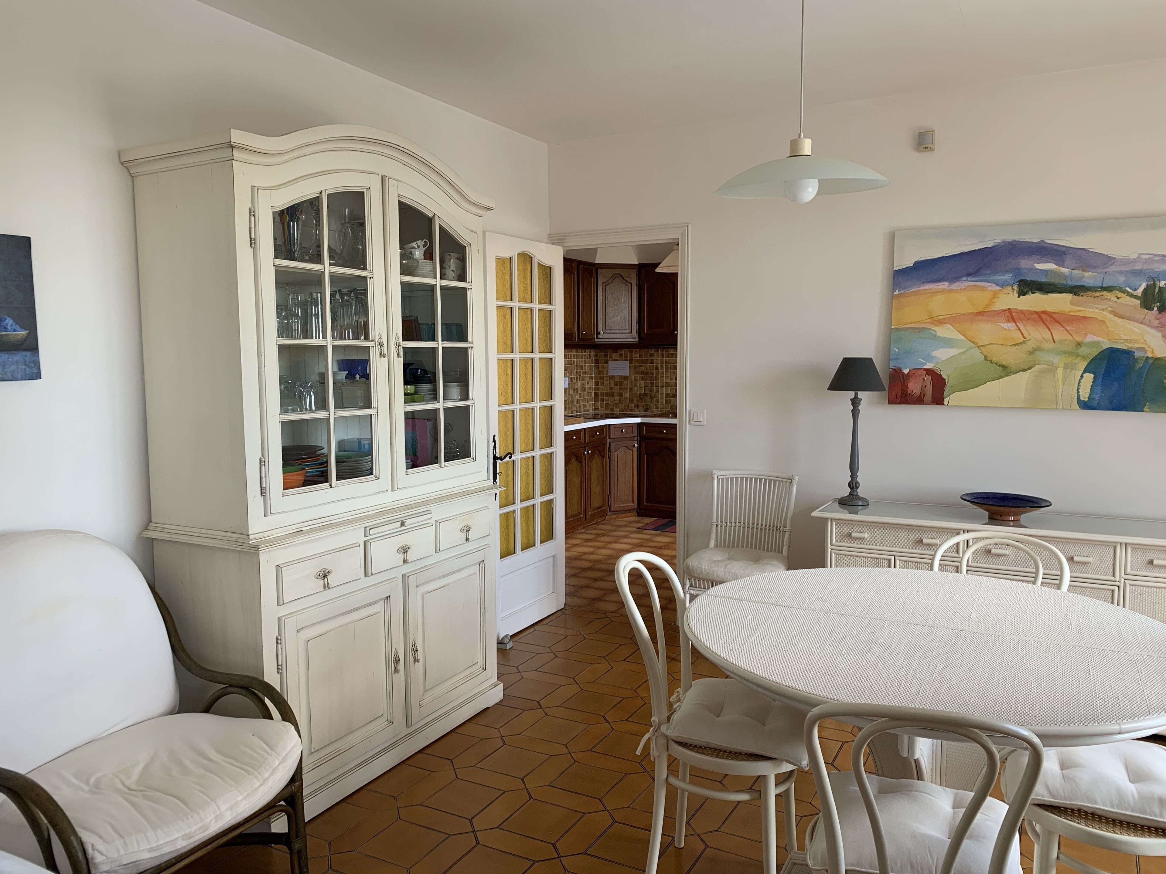 A dining area with a round table, white chairs, a cabinet with glass doors, and a painting on the wall.