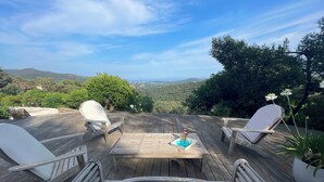 Um deck de madeira com uma mesa e quatro cadeiras, com vista para uma paisagem cênica com colinas e árvores.
