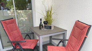 Una pequeña mesa exterior con dos sillas rojas acolchadas, una botella de champán y una planta en maceta.