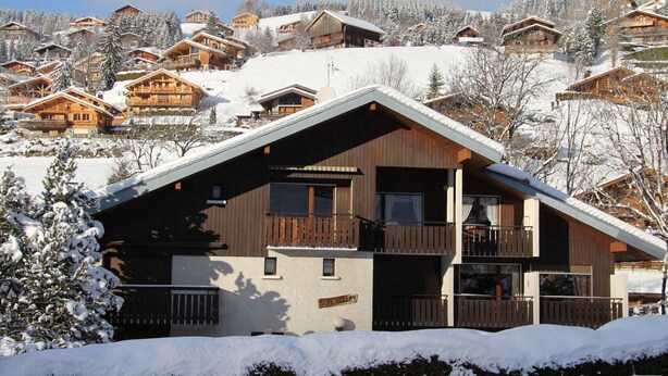 Una baita di legno con un balcone, circondata da alberi innevati e altre baite in zona montana.