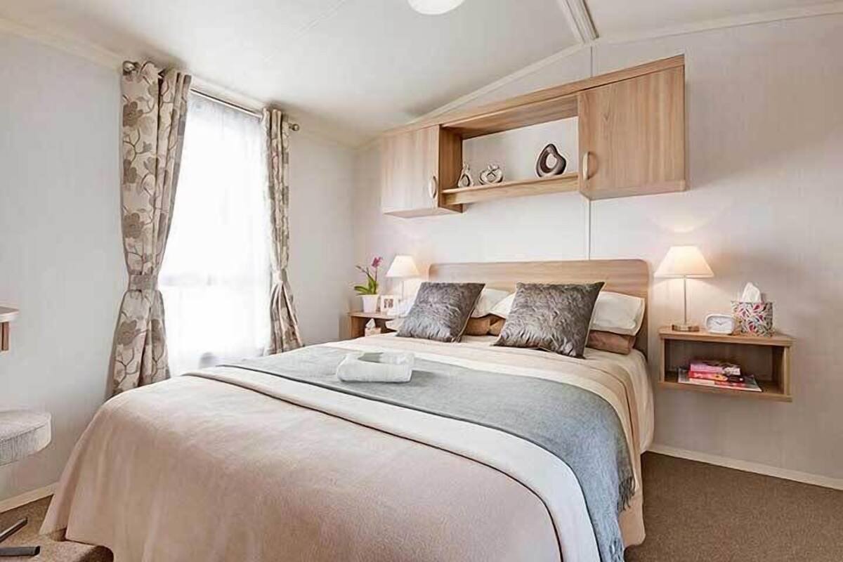 Une chambre à coucher bien rangée, avec un lit, des lampes de chevet, une étagère ornée d’objets décoratifs et une fenêtre avec des rideaux.