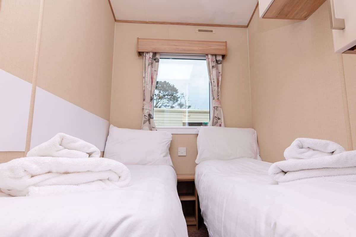 Une petite chambre accueillante, avec deux lits, chacun recouvert d’une couverture et d’un oreiller blancs, une fenêtre ornée de rideaux à motifs floraux et une vue sur les arbres à l’extérieur.