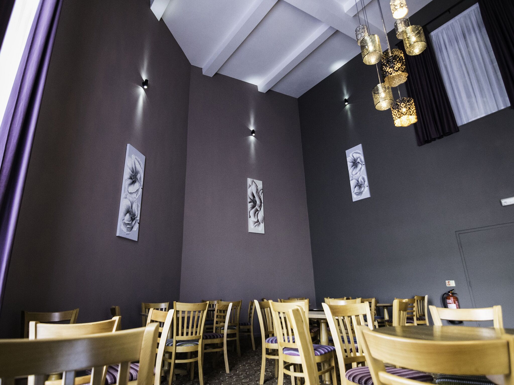 A restaurant interior with wooden tables and chairs, dark walls, and wall-mounted light fixtures.