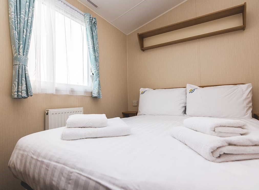 Ein ordentlich gemachtes Bett mit weißen Leinen und Handtüchern, ein Fenster mit durchsichtigen Vorhängen, ein hölzernes Regal über dem Bett und ein weißer Heizkörper unter dem Fenster.
