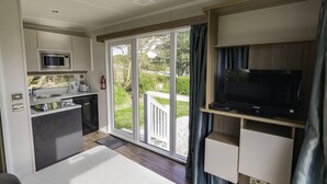 A compact kitchen with a microwave, toaster, and dishwasher, a flat-screen TV on a wooden stand, and a sliding glass door leading to a balcony. - Newquay Bay Holiday Resort (Newquay)