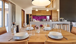 A dining area with a wooden table set for four, a kitchen in the background, and pendant lighting.