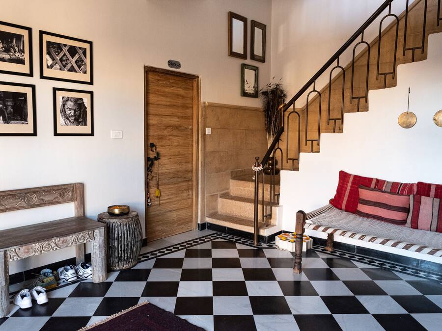 A room with a checkered floor, a wooden bench, a sofa with red cushions, a staircase, and framed pictures on the wall.