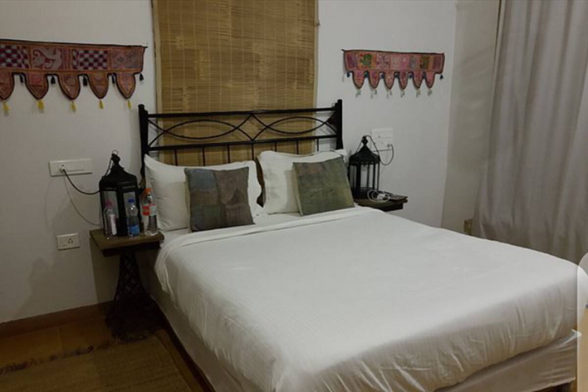 A neatly made bed with white linens, a wooden headboard, and decorative pillows. There is a nightstand with a lamp and a bottle of water. The room has a simple, clean design with a plain wall and a window with a curtain.