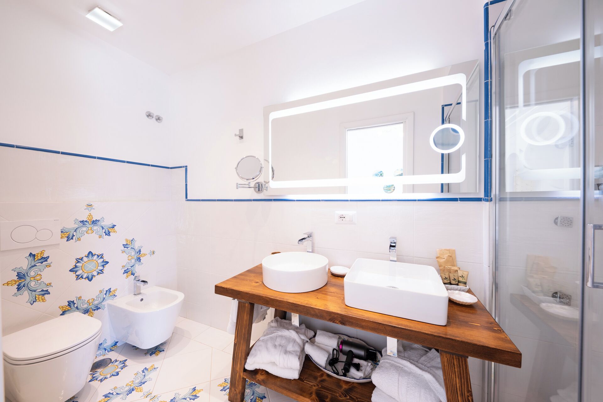 Et badeværelse med toilet, bidet, en trækommode med vask, et spejl og en brusekabine i glas.