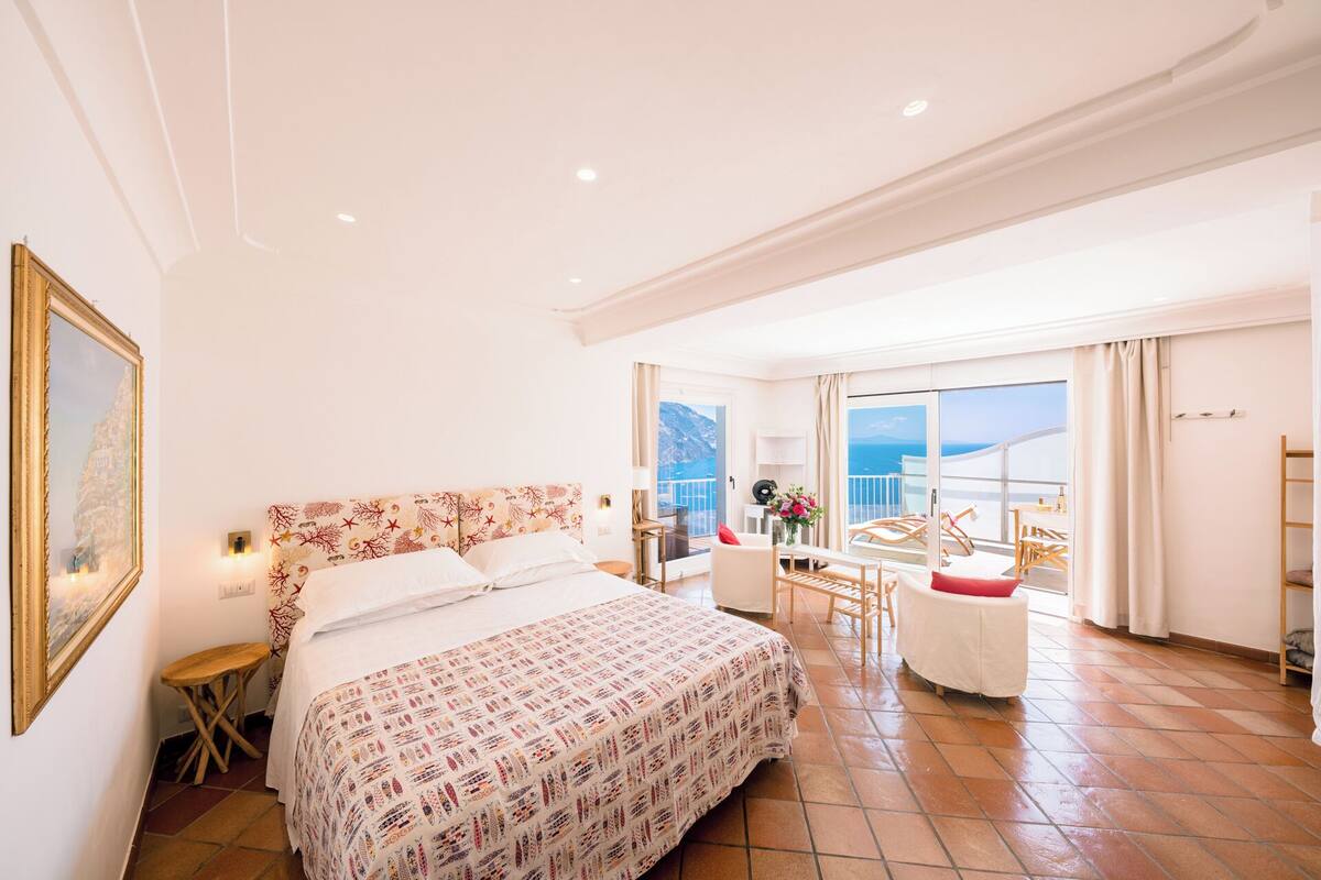 A spacious bedroom with a large bed, a balcony with a view of the sea, and a dining area with chairs and a table.
