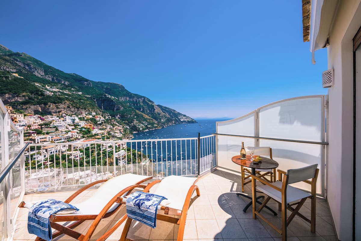 A balcony with a table and chairs, overlooking a coastal town and the sea.