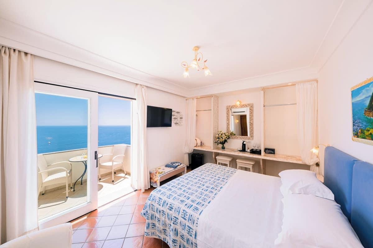 A hotel room with a bed, a desk, a chair, a TV, and a large window with a view of the sea.