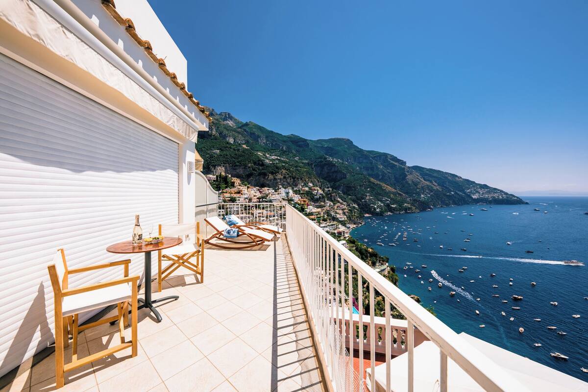 A balcony with a table and chairs, overlooking a coastal town and the sea.