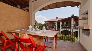 An outdoor patio with a wooden table set for dining, red chairs, and a view of a building with a tiled roof. - Appartamento Arianna (Manerba del Garda)