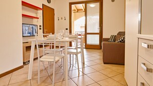 A dining area with a table and chairs, a television, and a sofa in a well-lit room with tiled flooring. - Appartamento Arianna (Manerba del Garda)