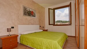 A bedroom with a bed, a nightstand, a bedside lamp, a window with a view, and a wall-mounted picture. - Appartamento Arianna (Manerba del Garda)