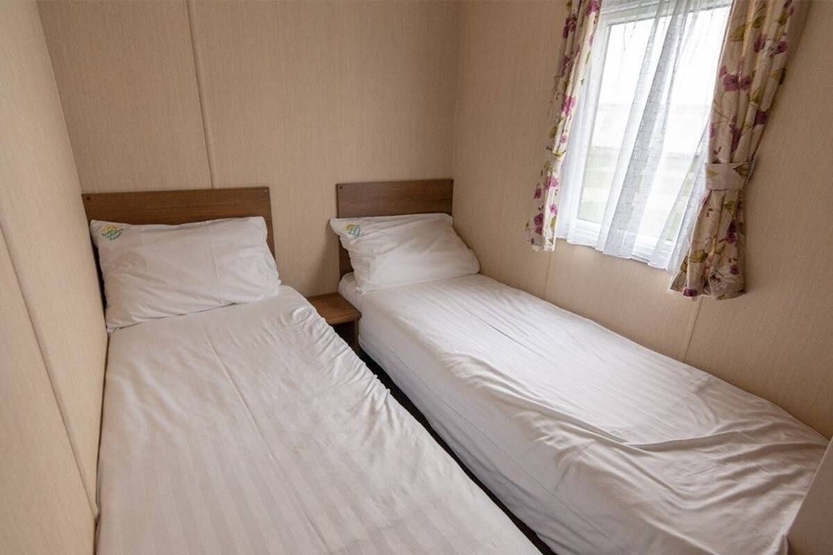 A small, single-room accommodation with two single beds, a wooden headboard, a window with floral curtains, and a plain wall.