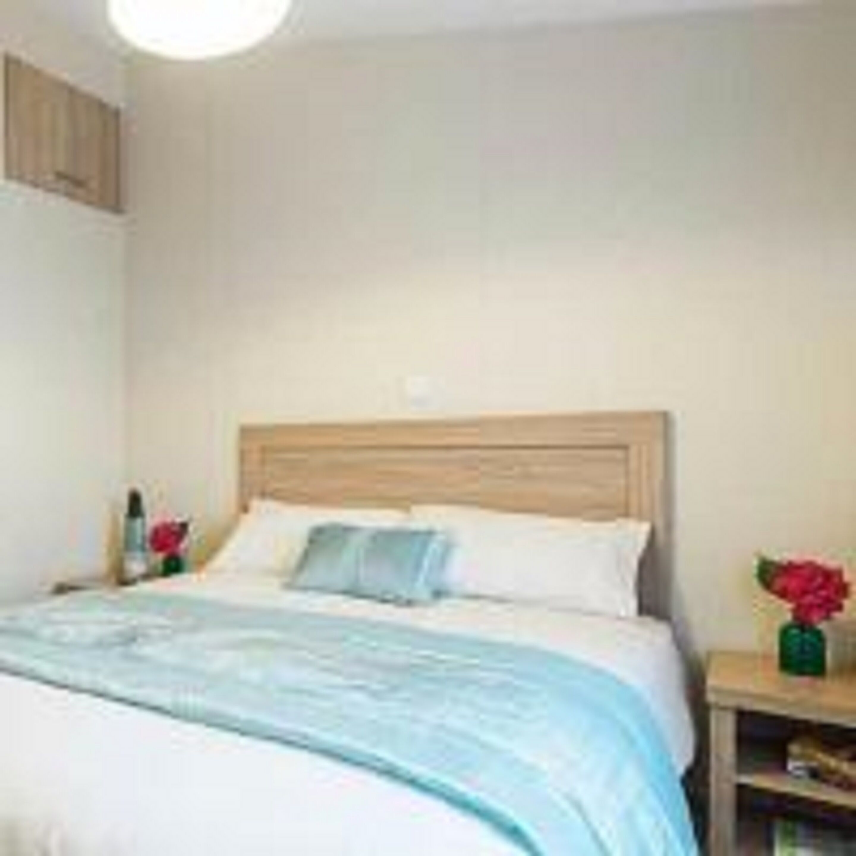 Une chambre à coucher avec une tête de lit en bois, un lit avec une literie blanche et bleue, une table de chevet ornée d’un vase rouge contenant une fleur, et une petite table sur laquelle est posé un vase vert.