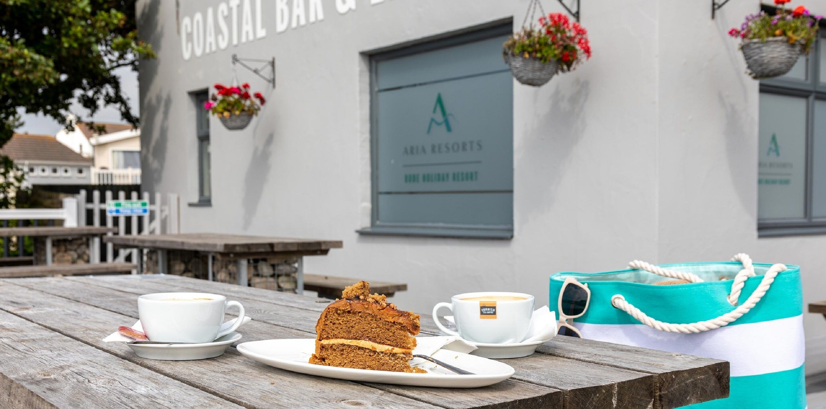 Una porción de pastel en un plato con una taza de café sobre una mesa de madera frente a un edificio con un letrero que dice 