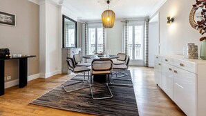 A dining area with a round table, four chairs, a black and white patterned rug, a hanging light fixture, and a white cabinet with decorative items.