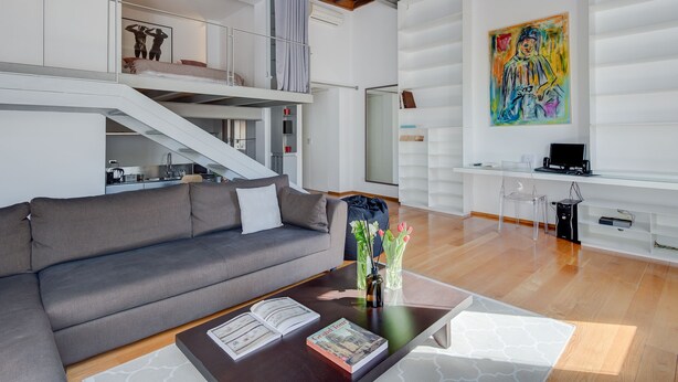 A spacious living area with a grey sofa, a coffee table with books and a vase, a staircase leading to an upper level, and a colorful abstract painting on the wall. - Trastevere Luxury Loft, unique, modern and with an Italian art gallery (Roma)