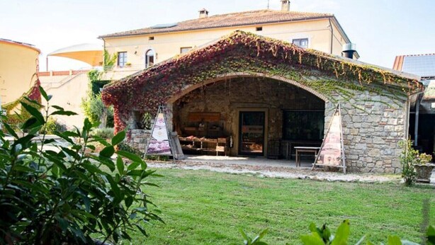 Un bâtiment en pierre doté d’un patio couvert, d’une cour gazonnée et d’un petit jardin avec des plantes au premier plan.