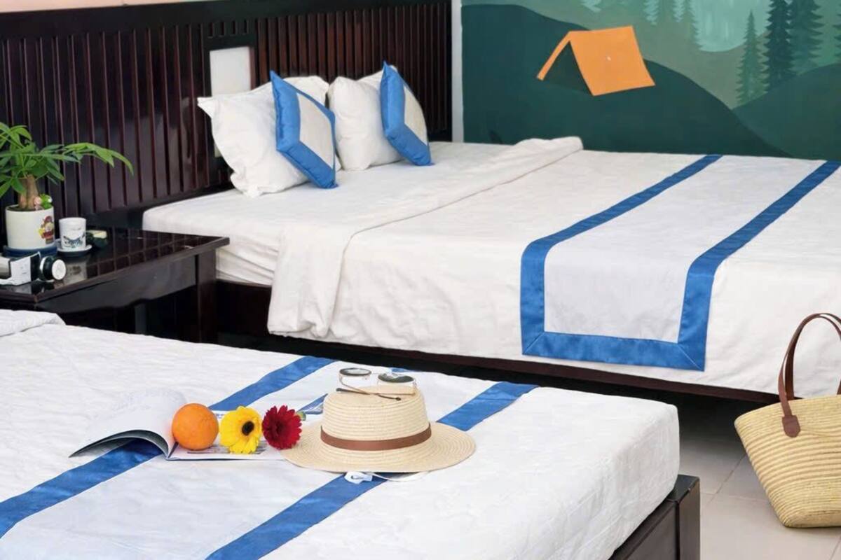 Habitación de hotel con dos camas, cada una con ropa blanca y detalles azules, un mural en la pared que representa un paisaje montañoso y una mesita con una planta y una taza.