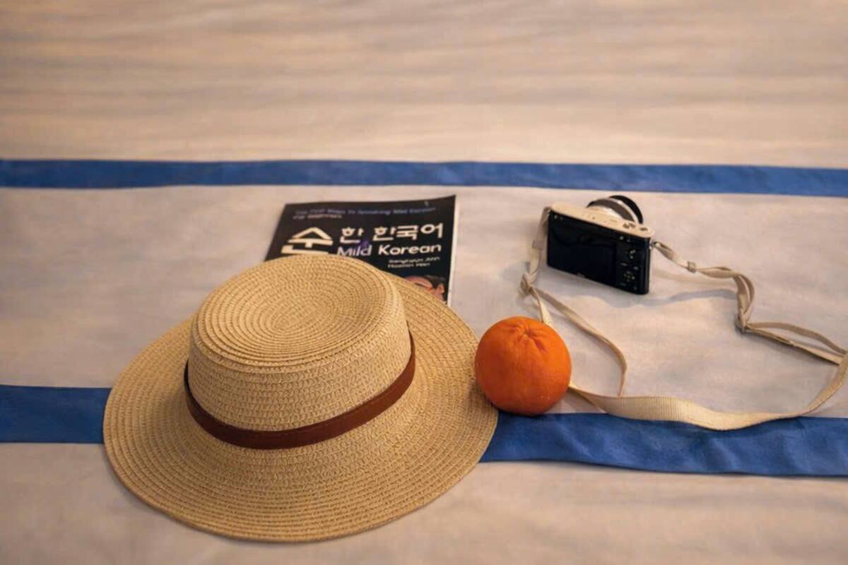 Una cama con cabecera a rayas, dos almohadas con diseños de diamantes azules, un sombrero de paja, una naranja y una cámara negra sobre un paño azul.
