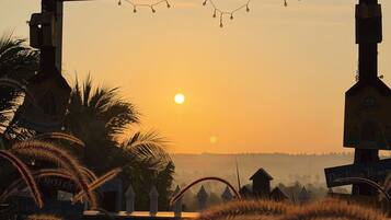 A sunset view framed by a wooden archway with string lights, palm trees, and a distant village.