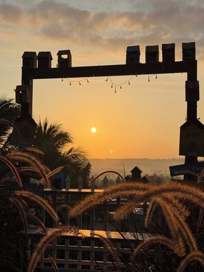 A sunset view framed by a wooden archway with string lights, palm trees, and a distant village.