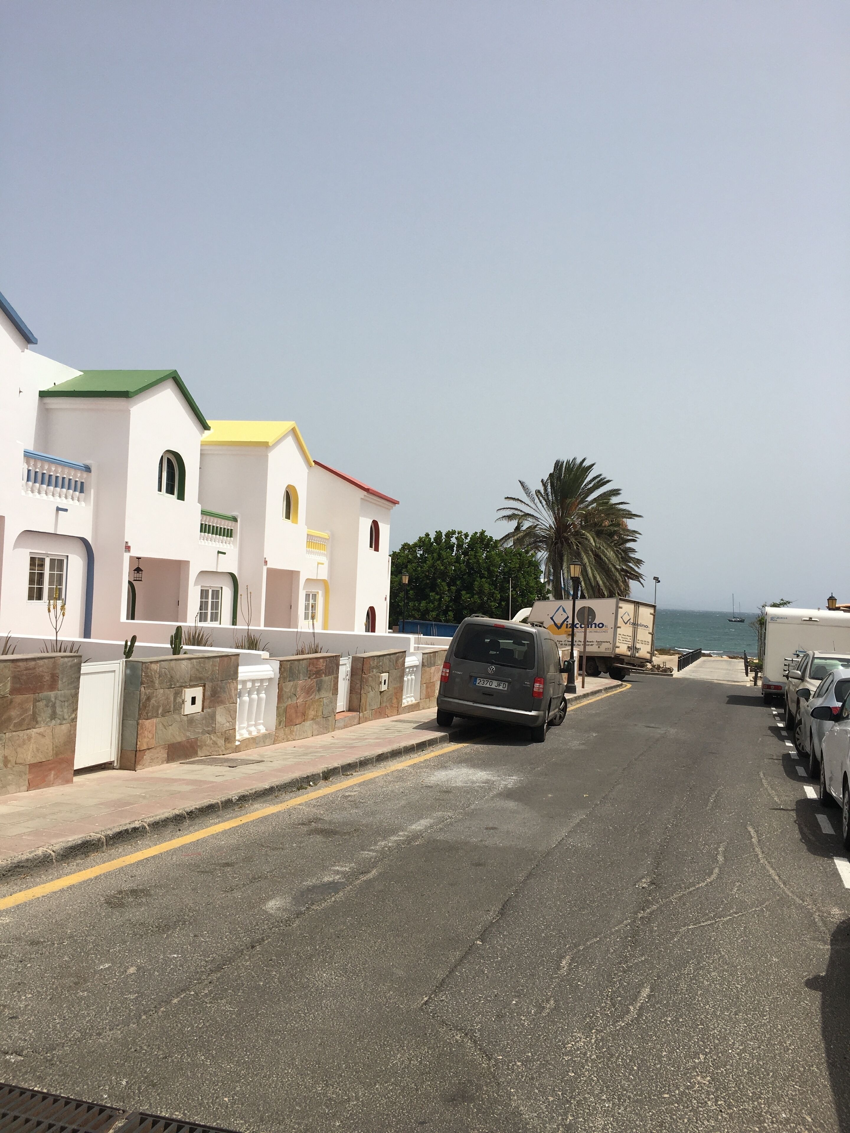 A coastal street with parked cars and a van, buildings with colorful facades, and a palm tree.
