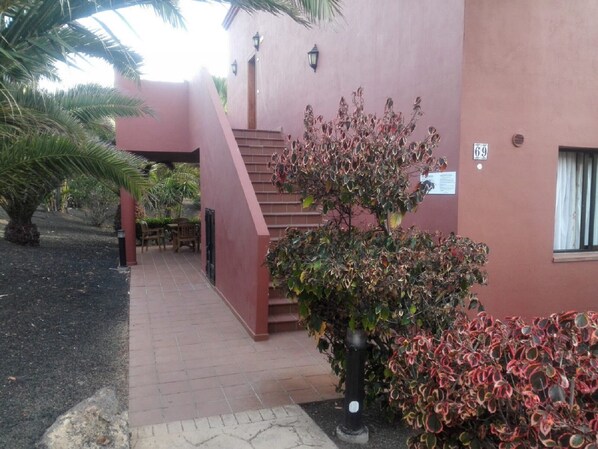 A building with a red exterior, a staircase, and a small garden area with a tree and shrubs.