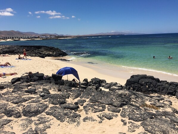 A beach with rocky foreground, clear blue water, and distant mountains.
