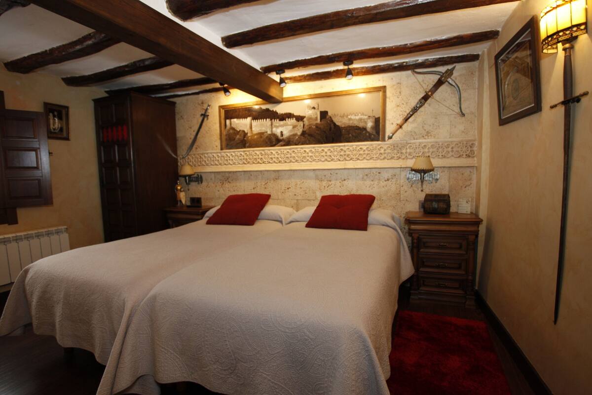 Uma cama de casal com almofadas vermelhas, uma cabeceira de madeira e um espelho acima. Há duas mesas de cabeceira com luminárias e um tapete vermelho no chão.