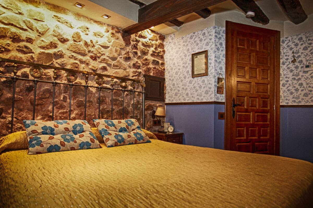 Quarto com parede de pedra, porta de madeira e cama com almofadas florais.
