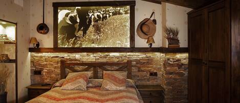 A bedroom with a stone wall, a bed with patterned bedding, a large framed picture, and a rustic wooden headboard.