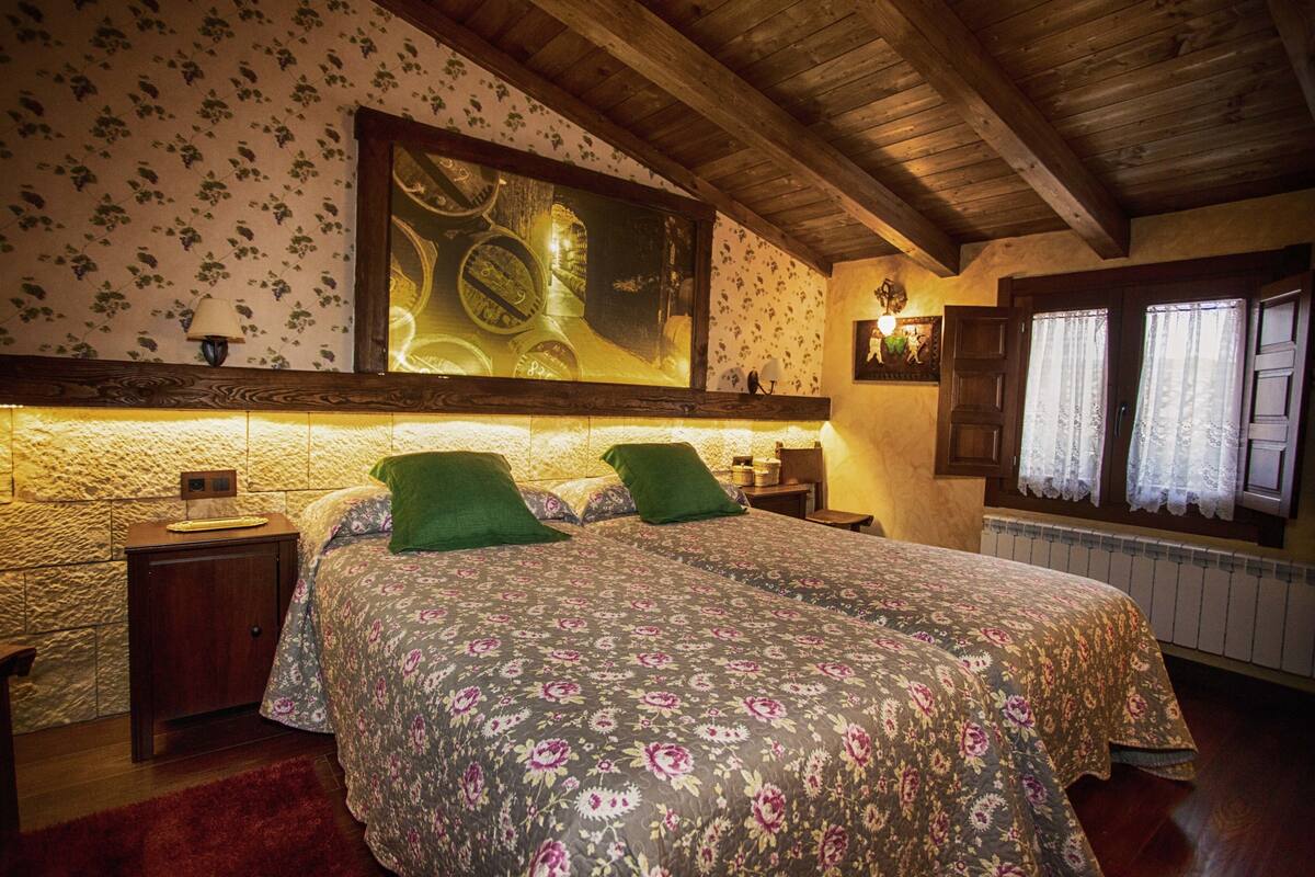 Quarto com teto de madeira, papel de parede floral, cama com almofadas verdes, mesa de cabeceira e uma grande moldura acima da cama.