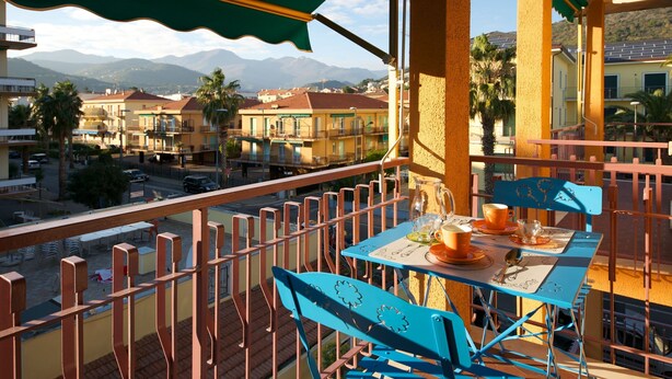 A balcony with a table set for two, overlooking a cityscape with buildings and mountains. - Fornaci 9 - 2-4 people 100 meters from the sea (Pietra Ligure)