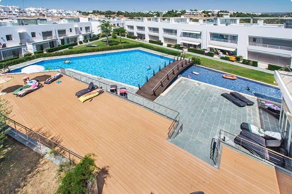 A rooftop pool area with sun loungers and a wooden deck, surrounded by modern buildings.