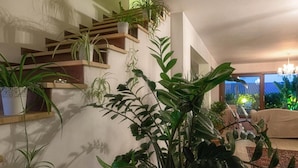 Una sala de estar con un jarrón grande de plantas verdes, estanterías de madera con plantas en macetas y vistas al exterior.