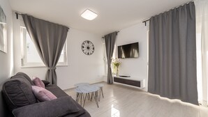 Ein modernes Wohnzimmer mit grauem Sofa, rundem Couchtisch, flachbildfernseher an der Wand und grauen Vorhängen.
