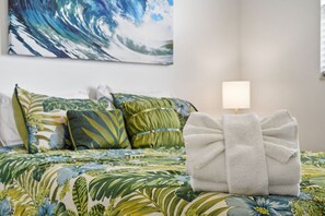Un lit avec une couvre-lit à motifs floraux, des oreillers ornés de motifs de feuilles et une serviette blanche sur laquelle est posée une petite lampe.