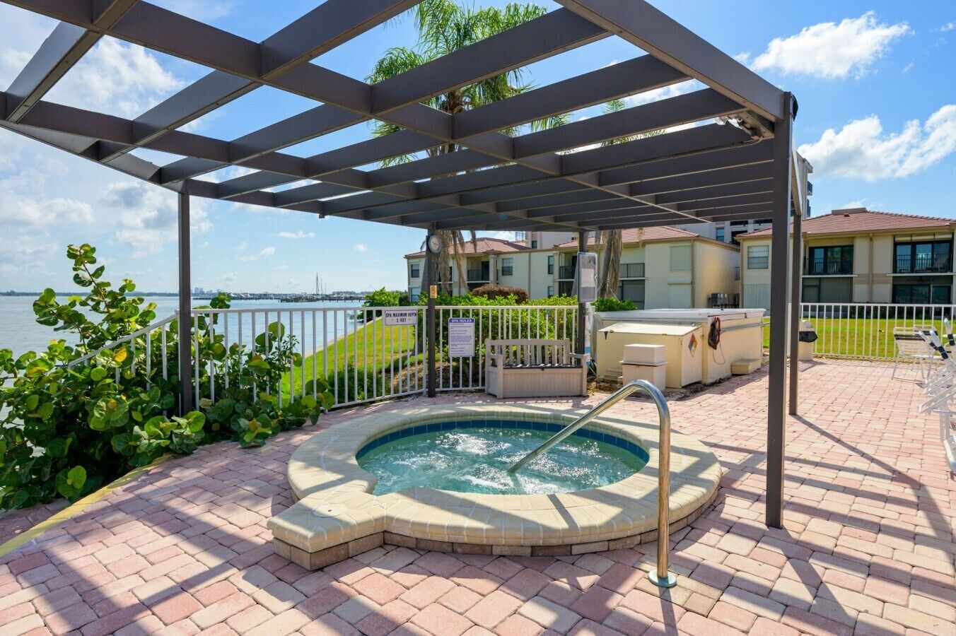 A covered outdoor swimming pool with a hot tub, surrounded by a brick patio, a white fence, and a view of a body of water and buildings.