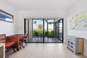 Une pièce avec une table et des chaises en bois, une porte-fenêtre coulissante donnant sur un balcon avec vue sur la mer et la verdure.