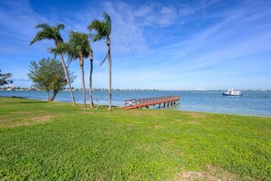 A grassy area with a wooden pier extending into a body of water, palm trees, and a distant boat. - St Pete Isla Casa C-103 2/2 Bath Amazing View! (Isla Del Sol)