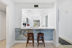 A modern kitchen with a marble island, two stools, and a view into the adjacent dining area. - St Pete Isla Casa C-103 2/2 Bath Amazing View! (Isla Del Sol)