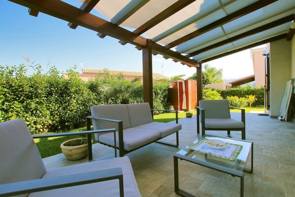An outdoor patio with a covered seating area, including a sofa and chairs, a table, and a potted plant.