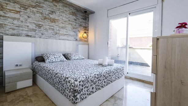Un dormitorio moderno con una cama grande, una mesita de noche, un cabecero de madera, una lámpara de pared y un ventanal corredero al exterior.